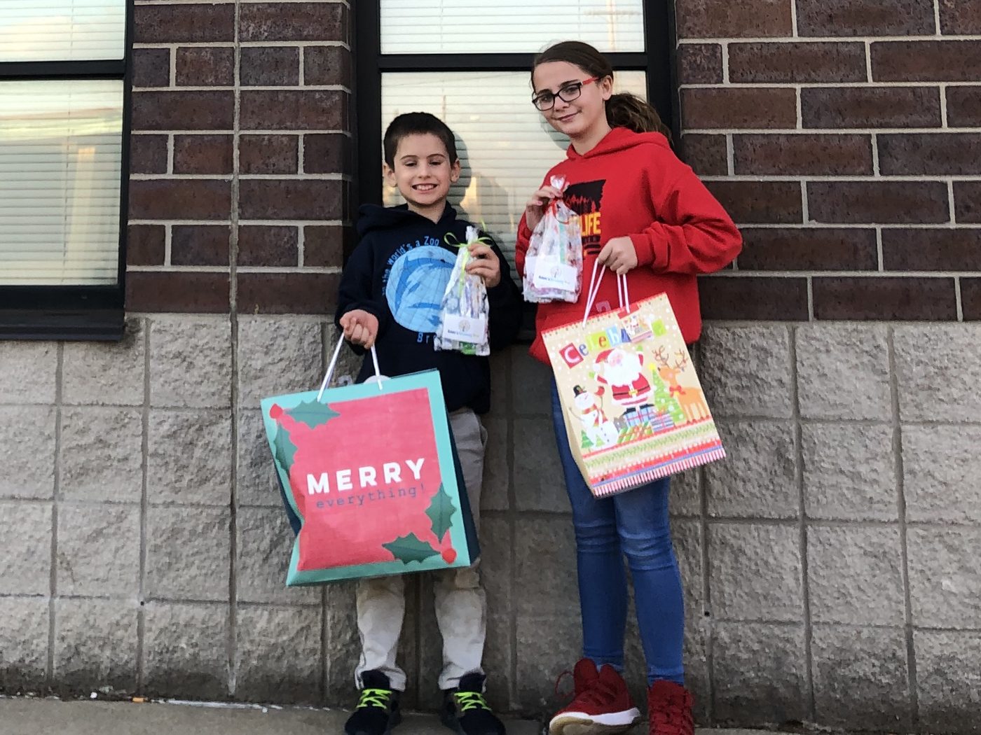 Two kids smiling and holding gift bags and small bags of dental supplies in front of a brick wall under the sign for The Center for the Homeless