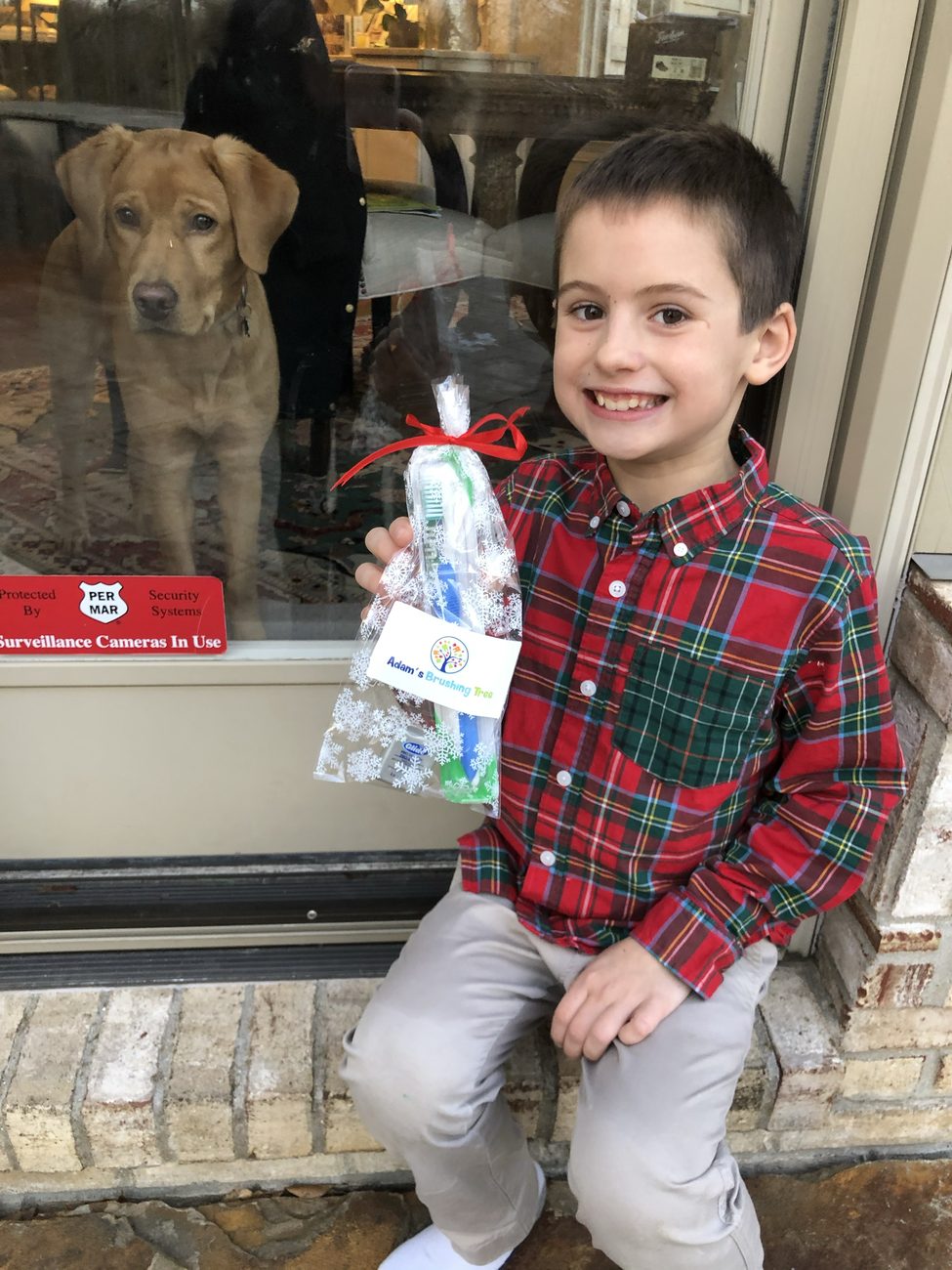 A boy smiling and holding a finished bag with a toothbrush, toothpaste and floss inside, tied with a red ribbon and a tag that says Adam's Brushing Tree