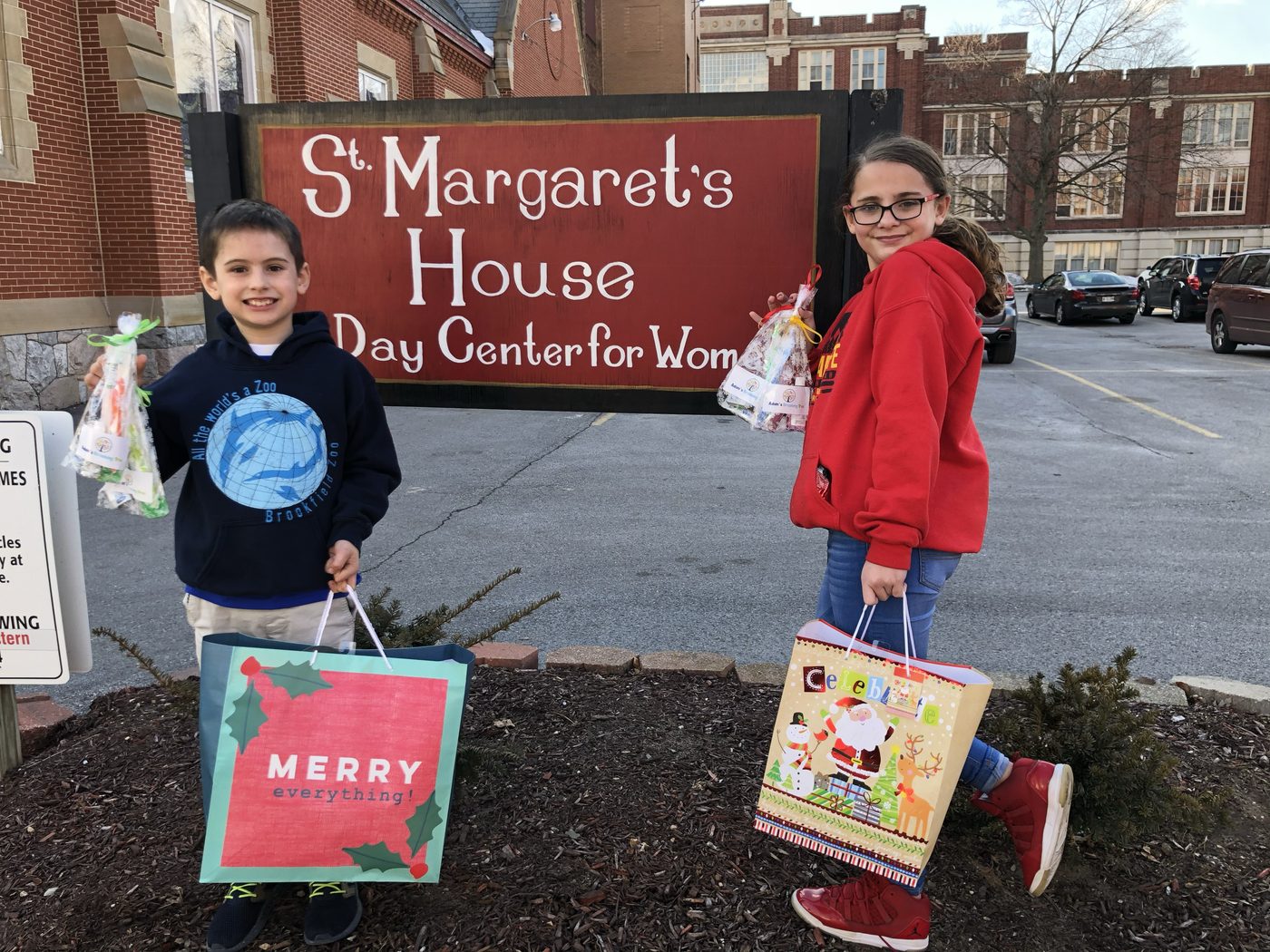 Two kids holding bags of dental supplies and large gift bags in front of the red St. Margaret's House day center sign
